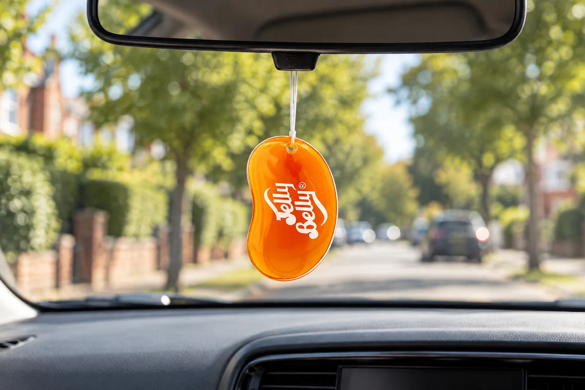 Jelly Belly Tangerine 3D air freshener hanging from a rear-view mirror over a tree lined street