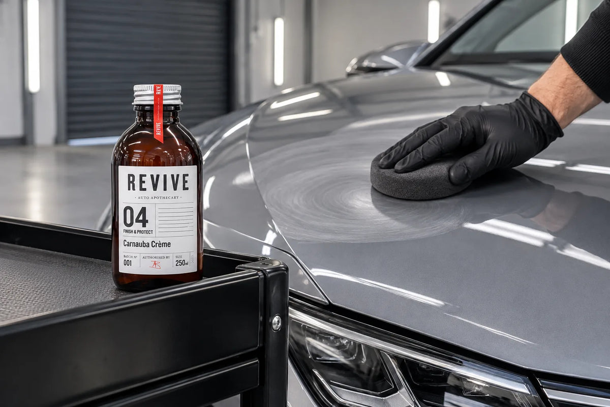 Revive Auto Apothecary 04 Carnauba Creme on a workshop trolley beside a grey car being waxed