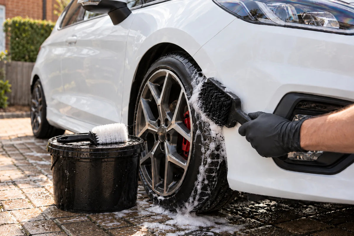 The Drivers HQ black tyre brush scrubbing the front tyre of a white Ford Fiesta ST with the white tyre brush resting on a wash bucket