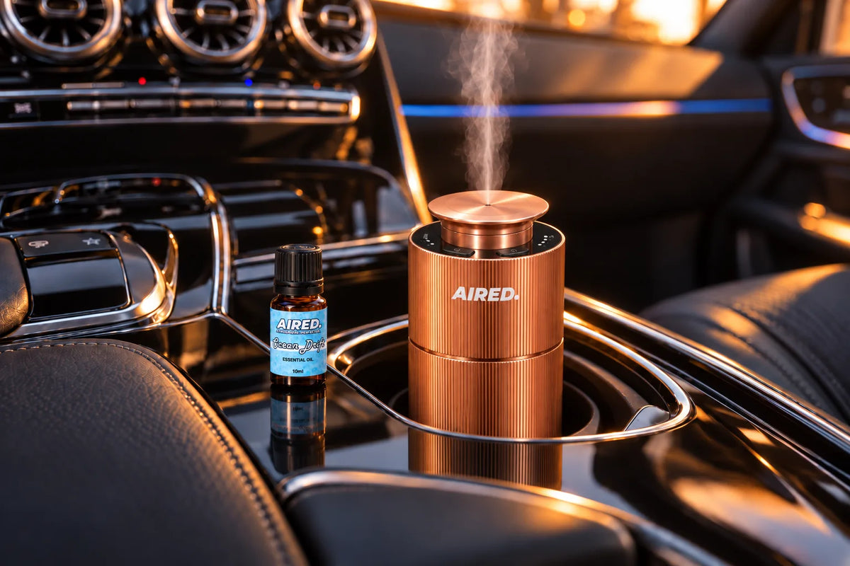 AIRED. Aura diffuser in rose gold misting in the front cup holder of a black Mercedes-Benz C-Class with the Ocean Drift essential oil beside it