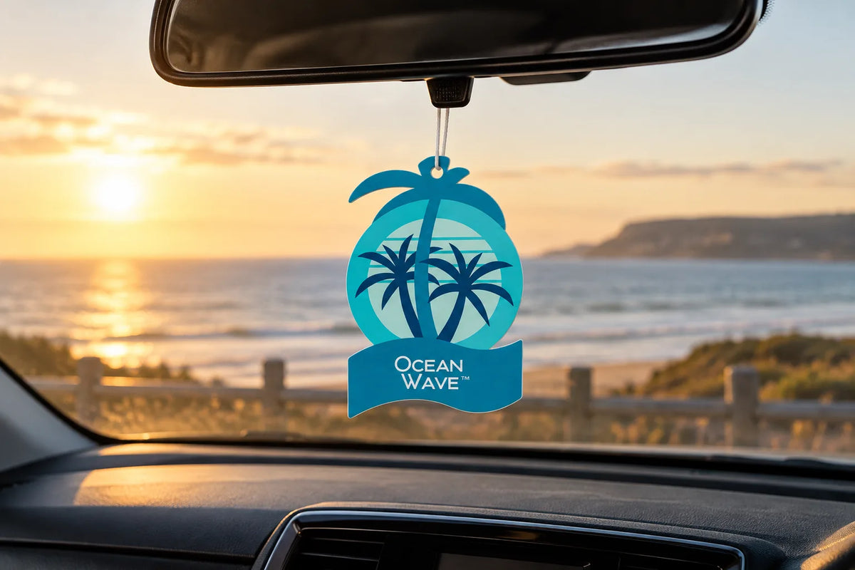 California Scents 2D Palm Ocean Wave air freshener hanging from a rear-view mirror with the sea at sunset behind