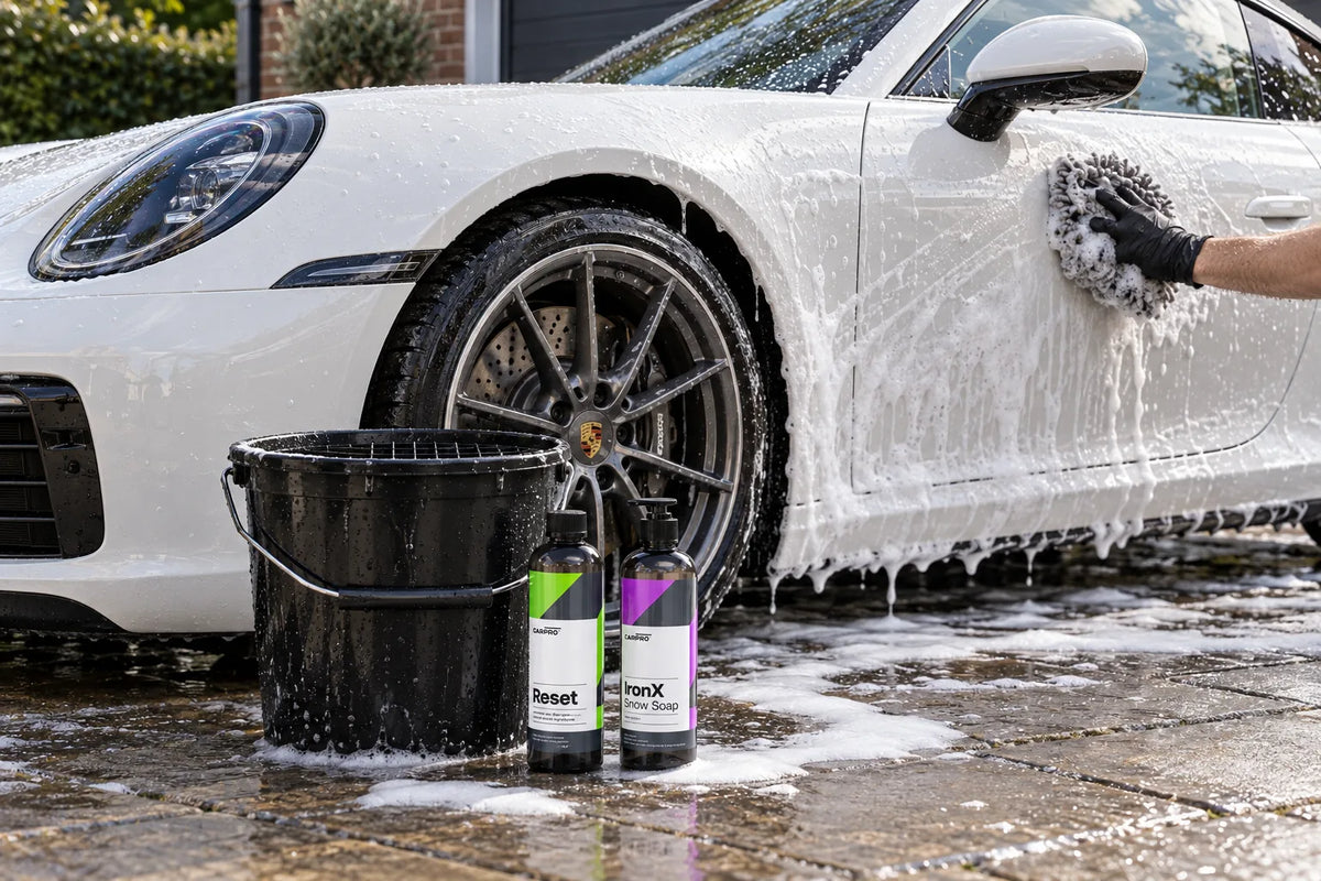 CARPRO Reset and IronX Snow Soap beside a wash bucket while a white Porsche 911 is washed