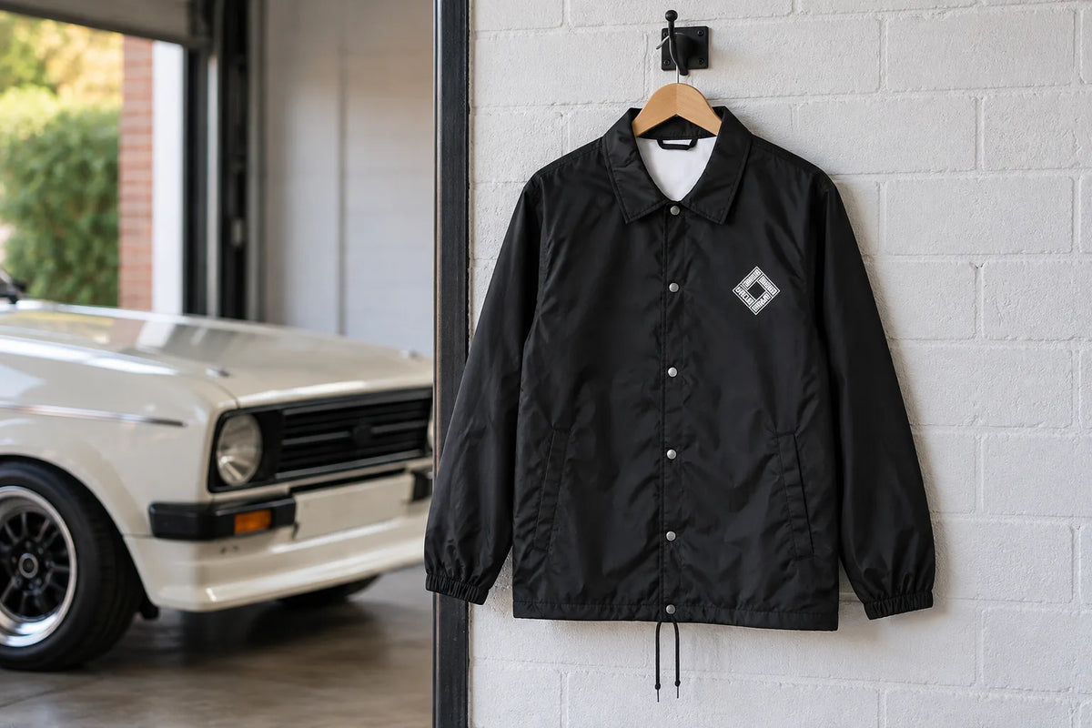 Black CDA coach jacket with the Crossed Dagger diamond chest logo on a hanger in a garage beside a white classic car