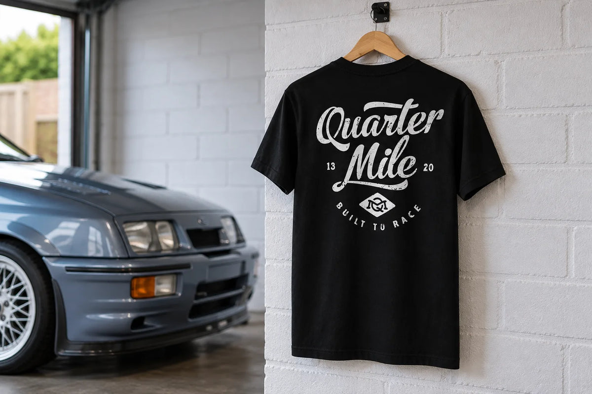 Black Quarter Mile Script tee showing its back print on a hanger in a garage beside a grey classic hatchback