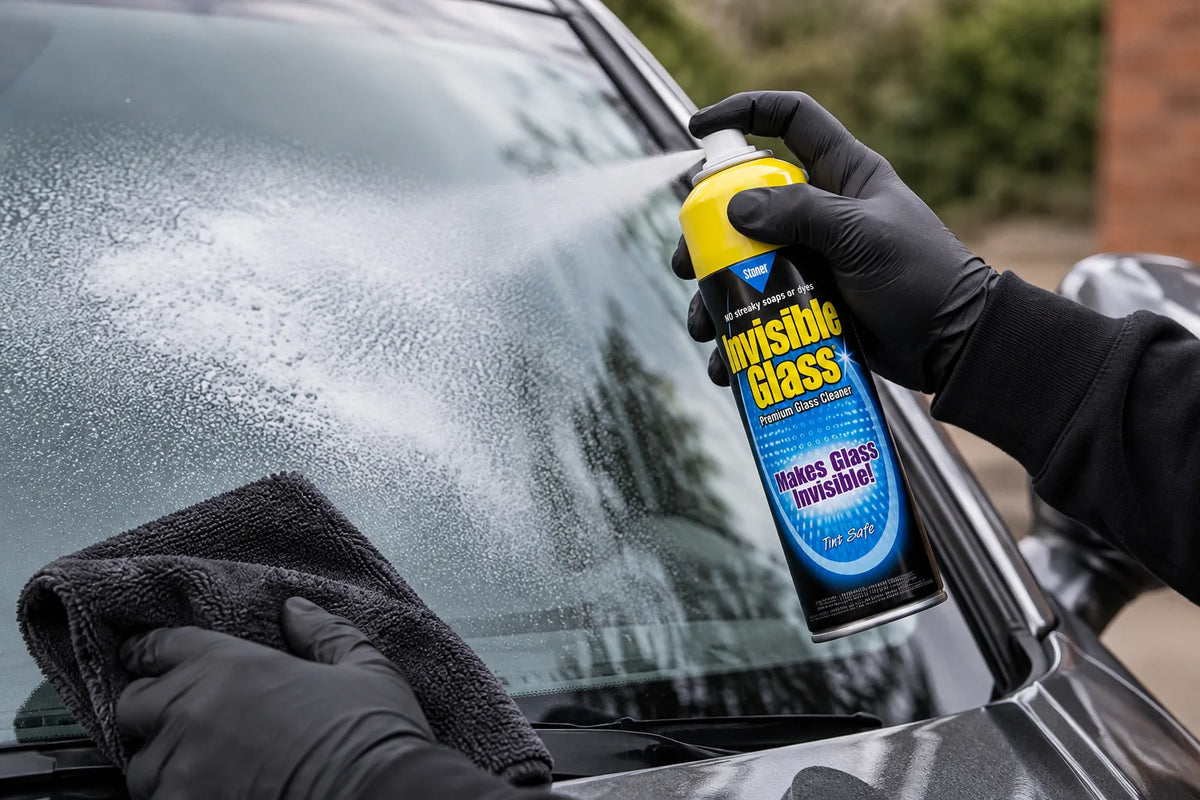 Stoner Invisible Glass aerosol being sprayed onto the windscreen of a grey car, a microfibre cloth ready to wipe