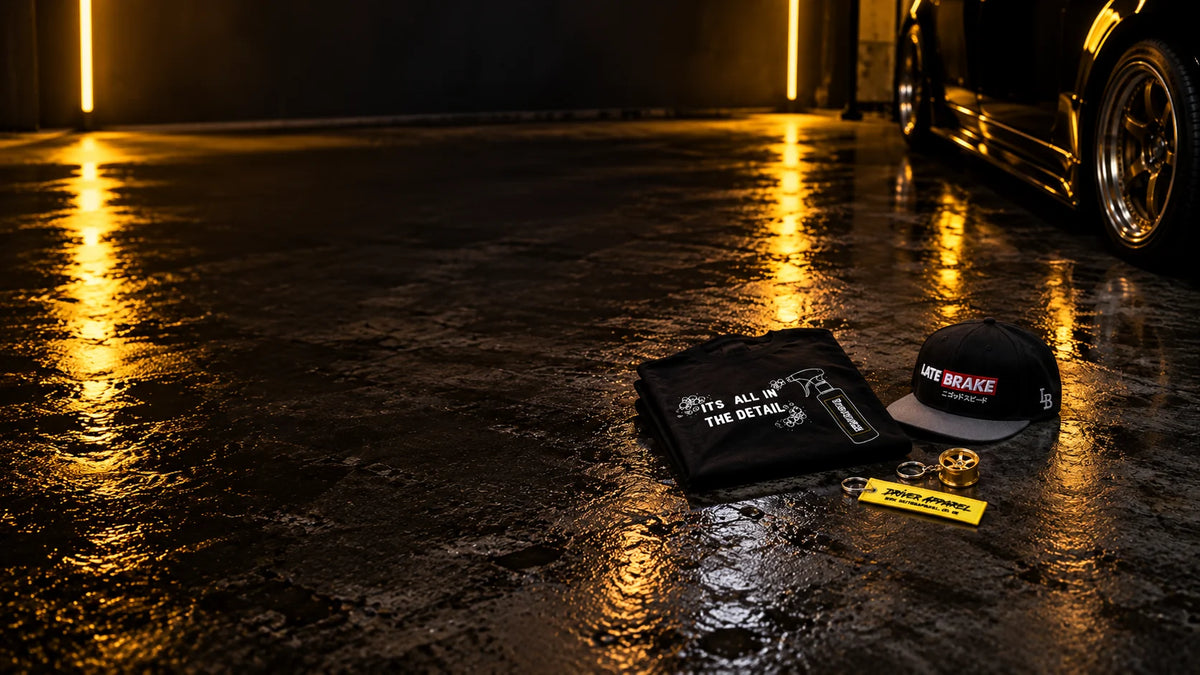 TDHQ Its All In The Detail t-shirt, a Late Brake snapback cap, a gold 6-spoke wheel keyring and a Driver Apparel flight tag keyring on a wet garage floor