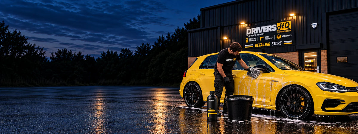 Detailer hand-washing a yellow hatchback with a FibreKing wash mitt, a bucket with a grit guard and AutoBead Pure Shampoo outside The Drivers HQ detailing store in Ilkeston at dusk