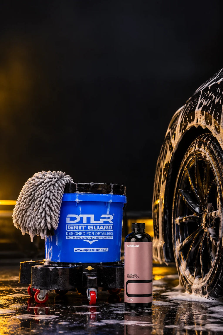 Safe wash kit: a DTLR Grit Guard bucket on its trolley with a microfibre wash mitt on the rim and AutoBead Ceramic Shampoo beside the foamed wheel of a black car