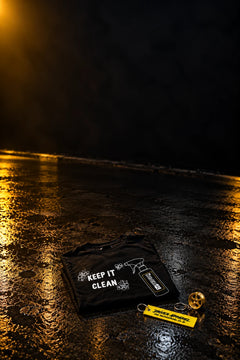 TDHQ Keep It Clean t-shirt folded on a wet garage floor with a Driver Apparel yellow flight tag keyring and a gold 6-spoke wheel keyring