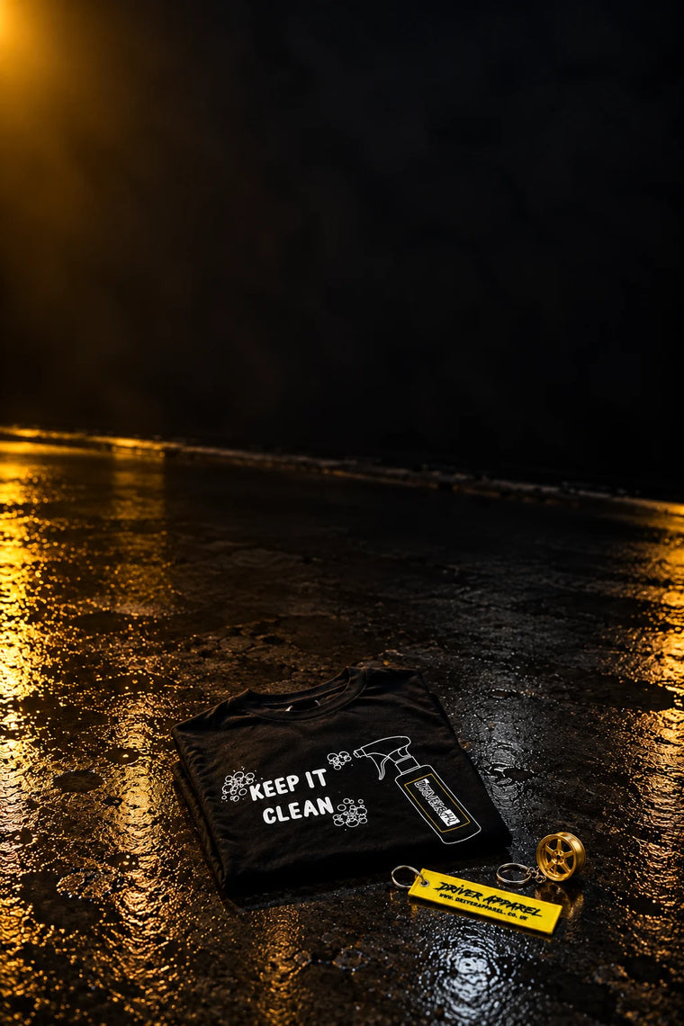 TDHQ Keep It Clean t-shirt folded on a wet garage floor with a Driver Apparel yellow flight tag keyring and a gold 6-spoke wheel keyring